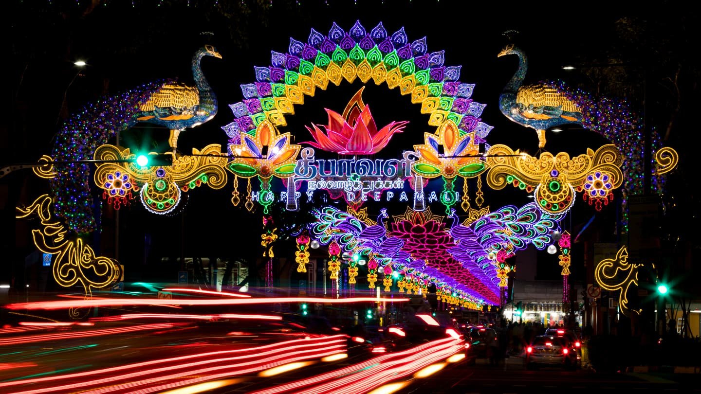 A festival arch of coloured lights over Serangoon Road at night: two peacocks and a lotus framed in bulbs above a greeting in Tamil and English, more arches glowing into the distance and the red trails of passing cars below.
