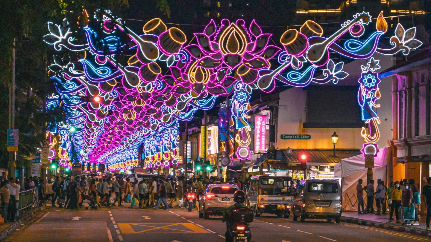Serangoon Road de nuit sous une voûte d'arches de néon rose, bleu et or en forme de lotus et d'instruments à cordes, une foule qui traverse au feu et la plaque verte de Campbell Lane à l'angle, à droite.