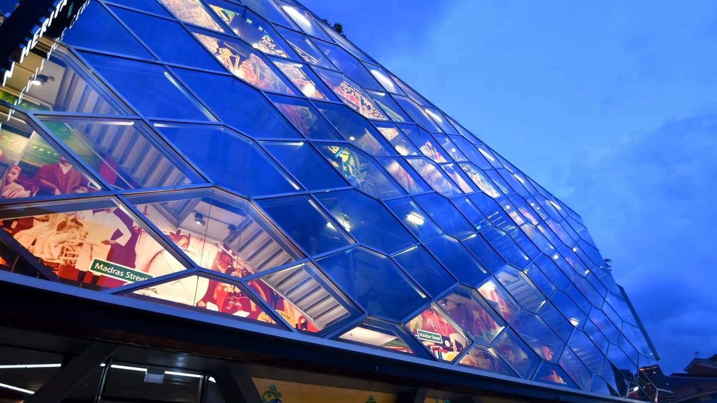 La façade de panneaux de verre inclinés de l'Indian Heritage Centre, éclairée de bleu au crépuscule, avec des fresques peintes visibles derrière la vitre et une plaque de Madras Street sur les vitres du bas.