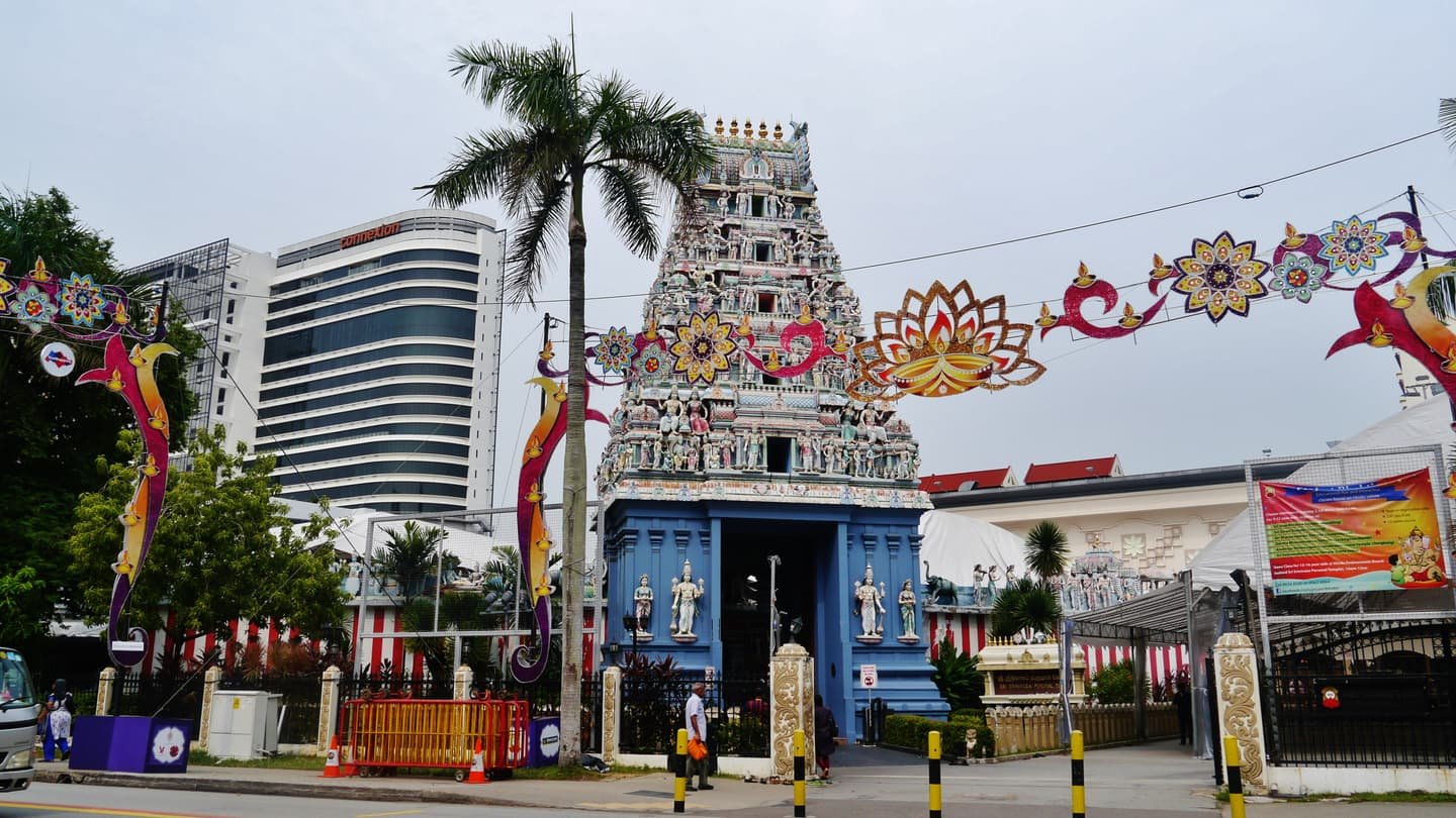 Le haut gopuram bleu et blanc du temple Sri Srinivasa Perumal, couvert de figures peintes de divinités, derrière les grilles du temple sur Serangoon Road, avec des décorations de fête en forme de lotus et d'oiseaux tendues au-dessus de la rue.