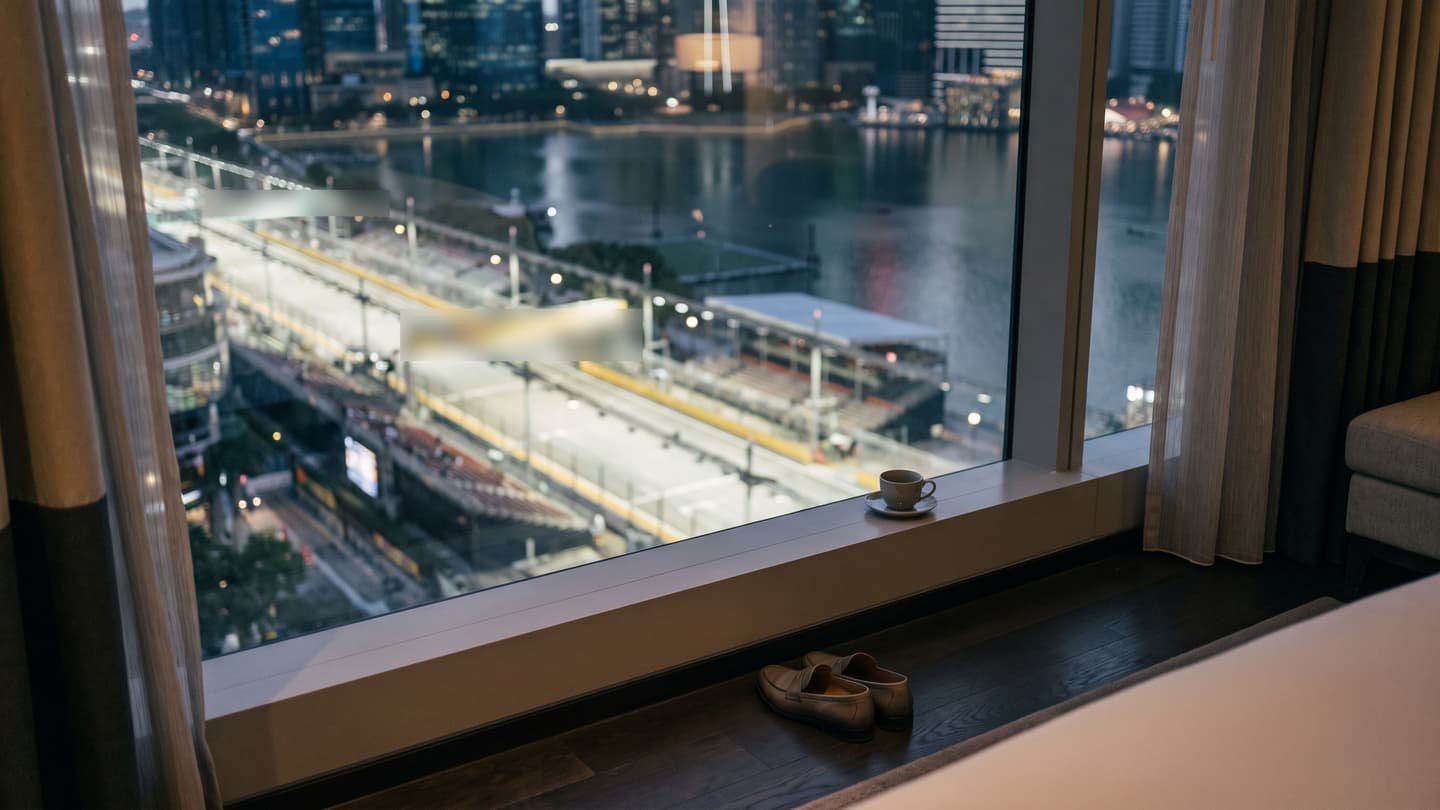 A hotel room at night, a cup on the sill and a pair of loafers on the floor, the window looking straight down onto the floodlit barriers and grandstands of the street circuit beside the bay.