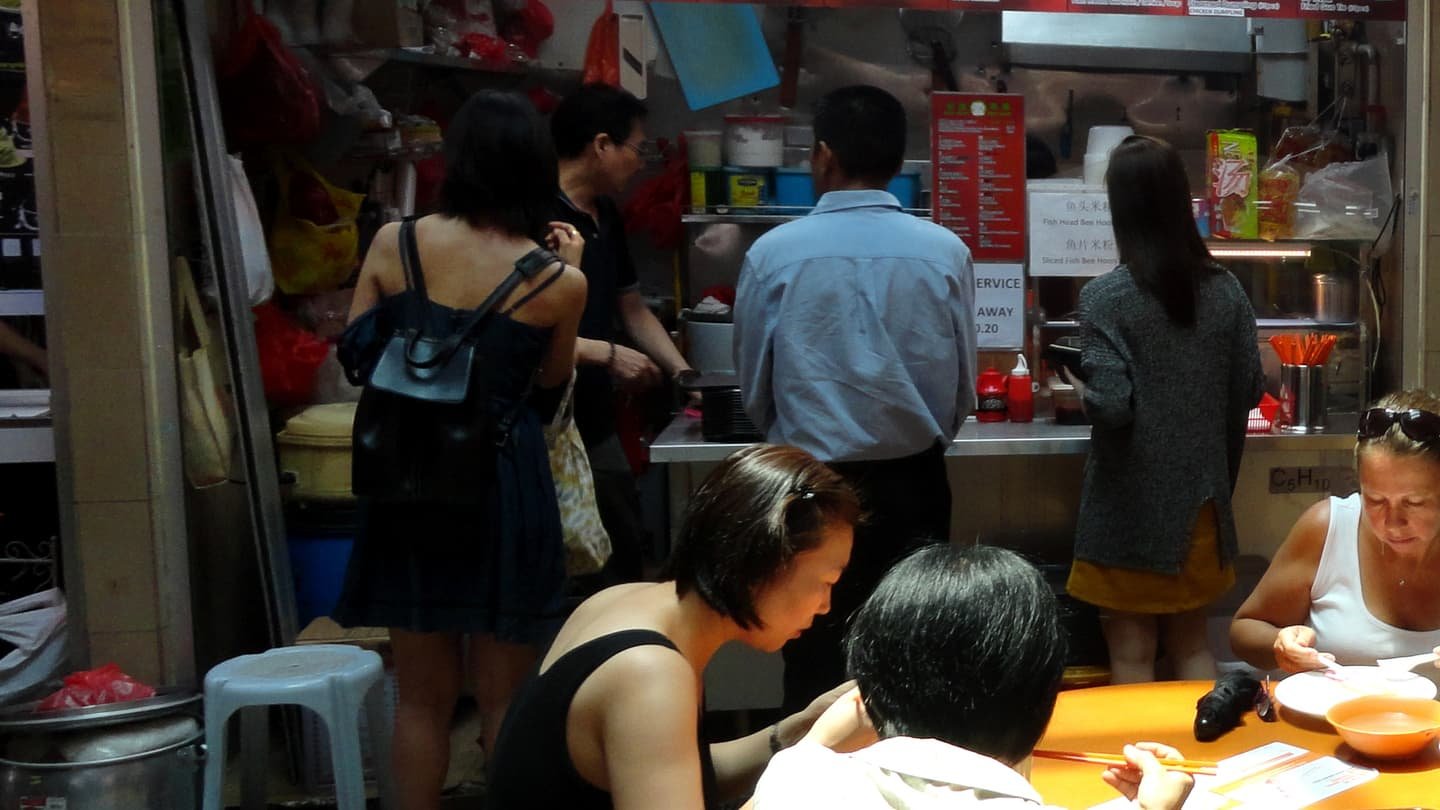Le comptoir d'un stand de hawker centre, une file de gens en tenue décontractée attendant sous une carte imprimée en rouge et, au premier plan, des convives penchés sur leurs bols autour d'une table ronde orange.