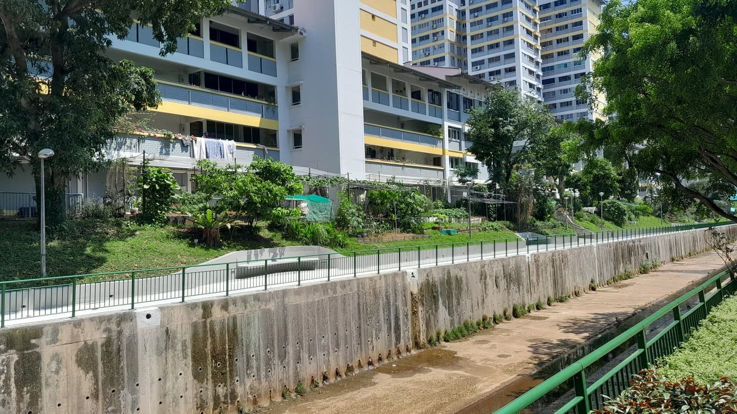 Des barres de logement public derrière un jardin communautaire de petites parcelles, du linge étendu sur des perches depuis une coursive et un large canal de béton bordé de garde-corps verts au premier plan.