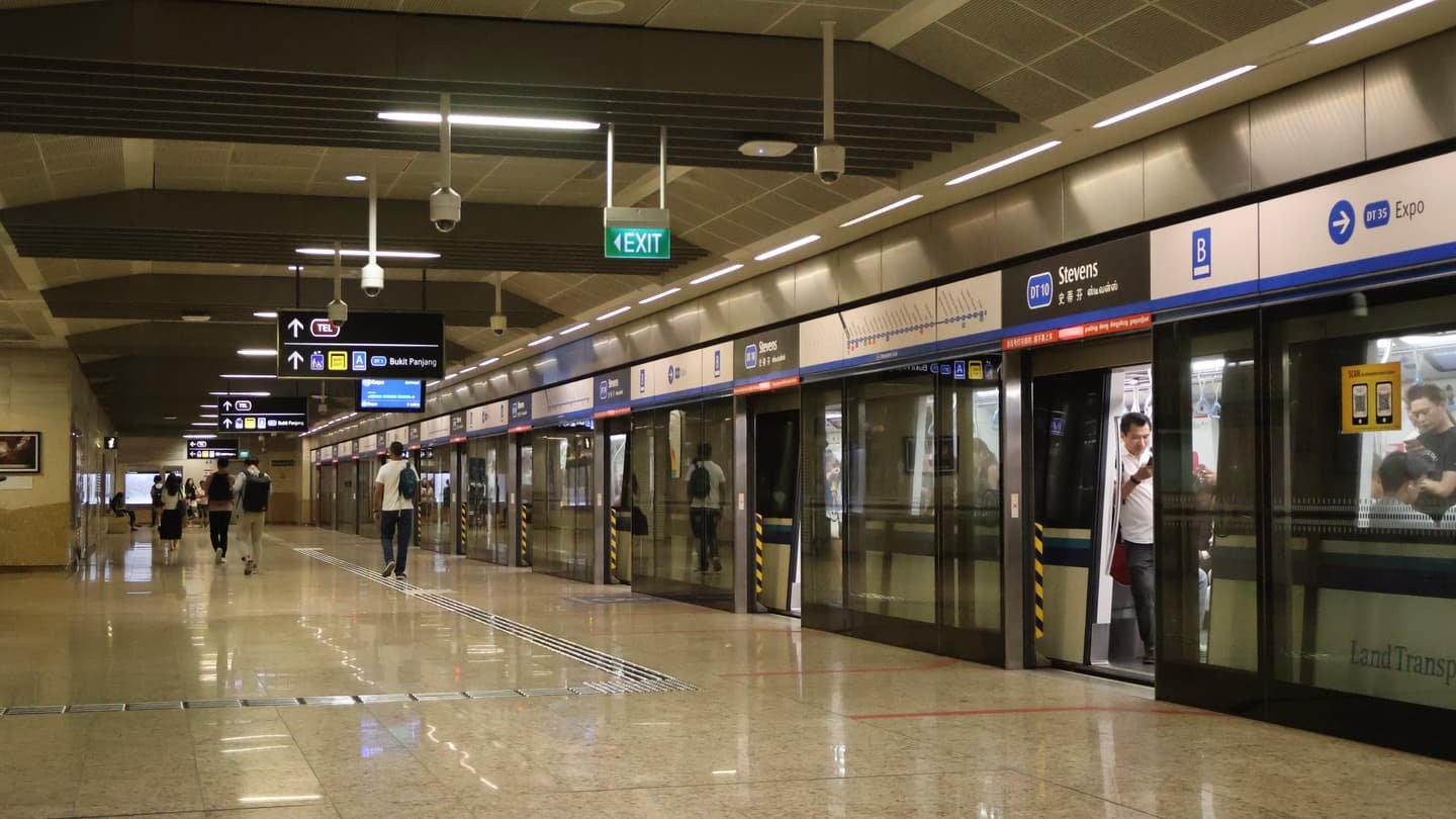 Un quai de MRT souterrain très éclairé, avec des façades de quai vitrées toute hauteur, une rame arrêtée portes ouvertes et quelques voyageurs marchant sur le sol poli.