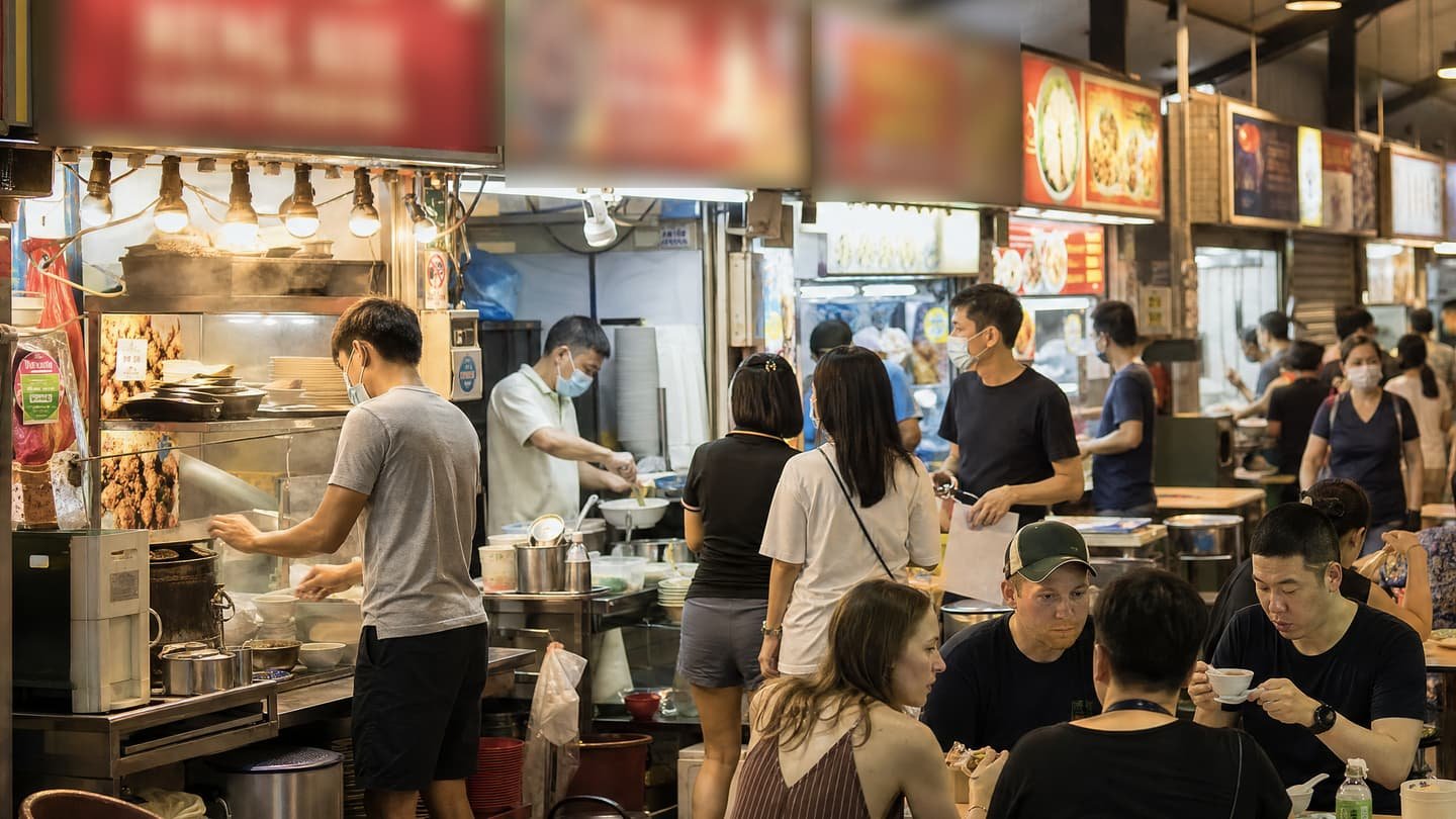 A busy hawker centre at mealtime: lit stall signboards overhead, a cook plating at the counter, and locals and visitors sharing the tables in front.