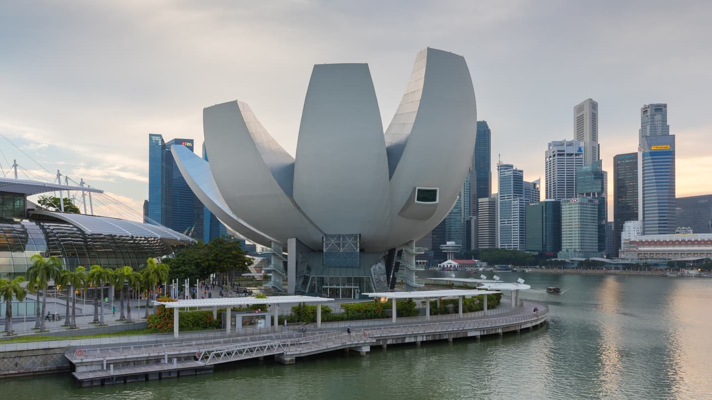 El ArtScience Museum, blanco y con forma de flor de loto, junto al agua de Marina Bay con luz suave de mañana, el paseo curvándose delante y las torres del distrito financiero al otro lado.