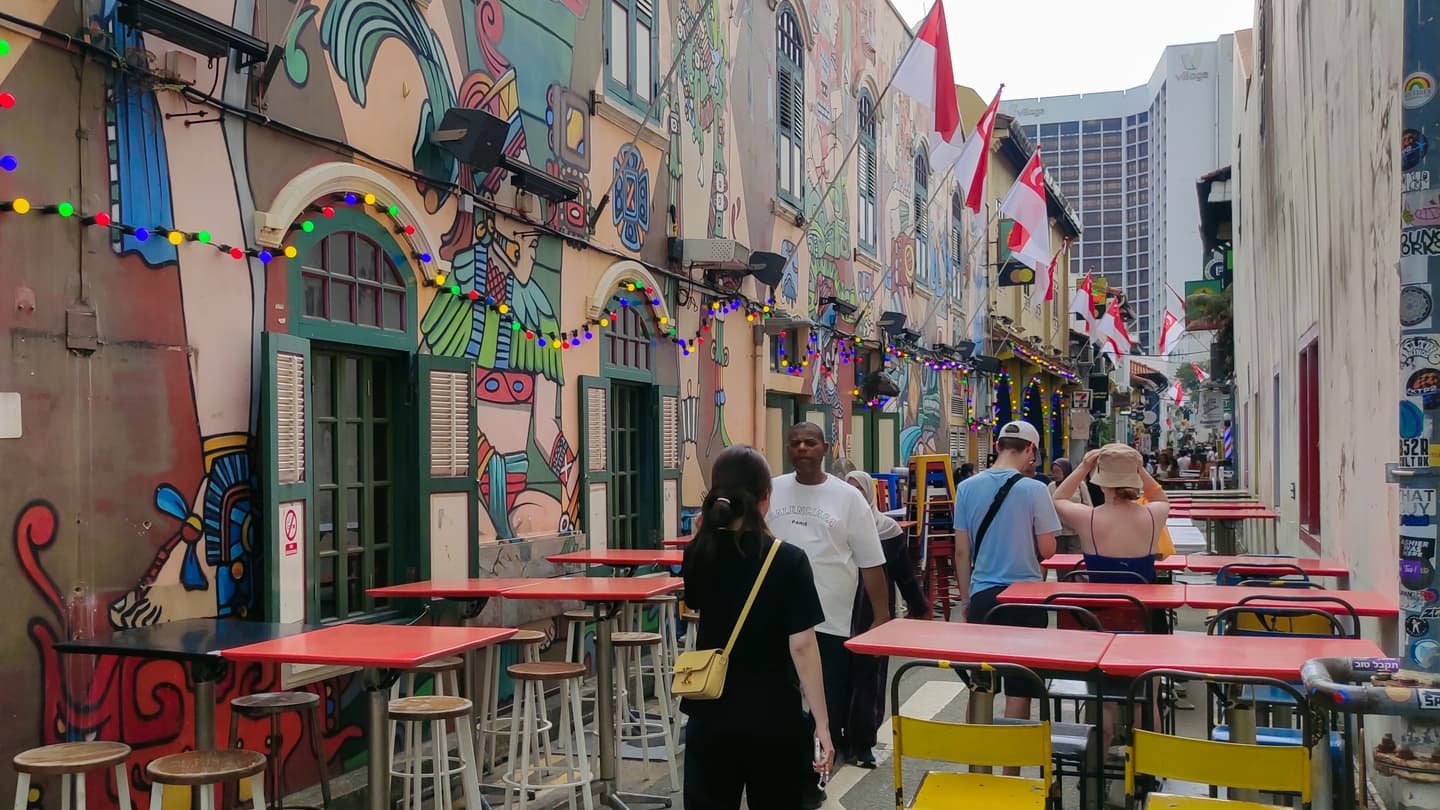 Un callejón estrecho de casas-tienda bajas pintadas con murales vivos, guirnaldas de luces de colores y banderitas en lo alto, mesas rojas de café en la acera y visitantes paseando entre ellas.