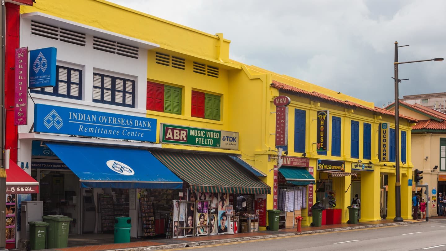 Una hilera de casas-tienda de dos plantas en Serangoon Road pintadas de amarillo y blanco, con contraventanas azules y rojas, toldos de rayas y carteles colgados sobre el pasaje cubierto.